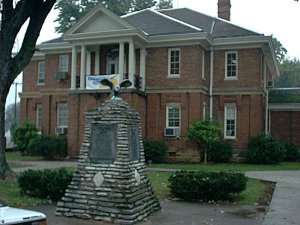 Trimble County Courthouse, a red brick building, sits under a dreary sky with an old stone plaque and a statue. 